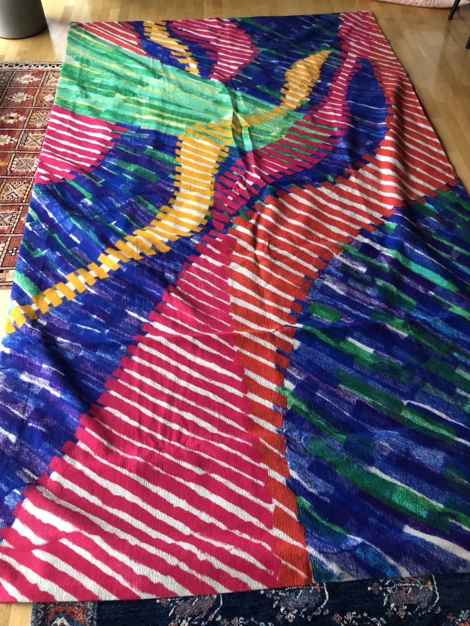 Fotografi av et stort billedteppe lagt utover et gulv. Diagonale striper i rosa, blått, grønt, gult og oransje møtes i brede, buede baner.