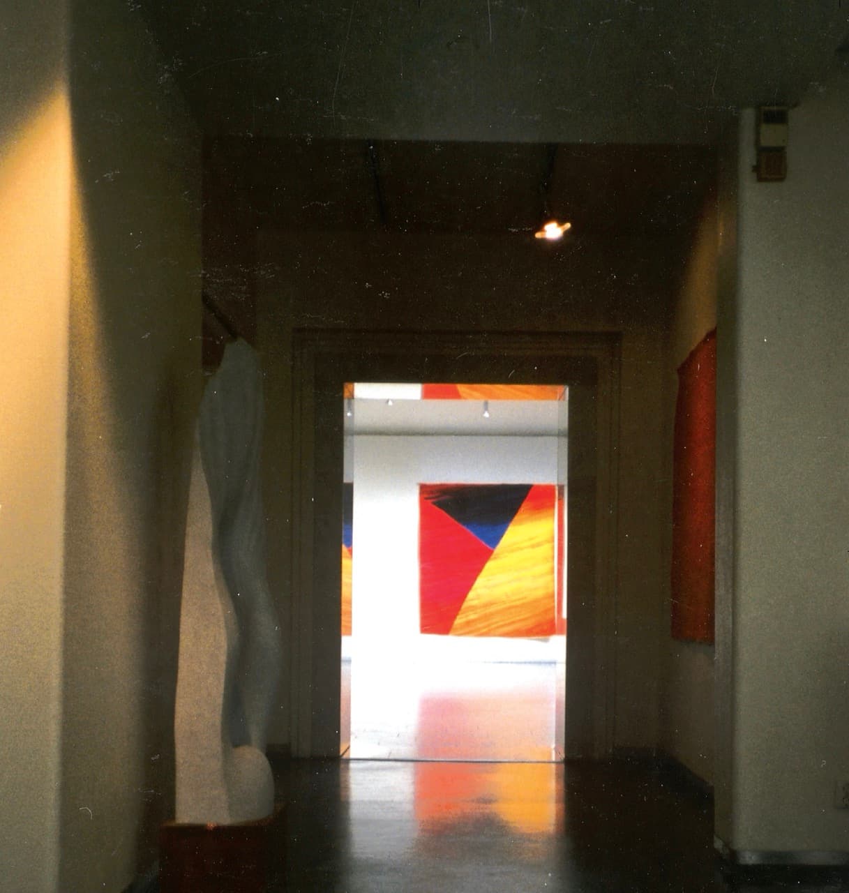 Fotografi fra et galleri. Gjennom en mørk døråpning ses et billedteppe i rødt, blått og oransje i et opplyst rom, med en lys skulptur til venstre.