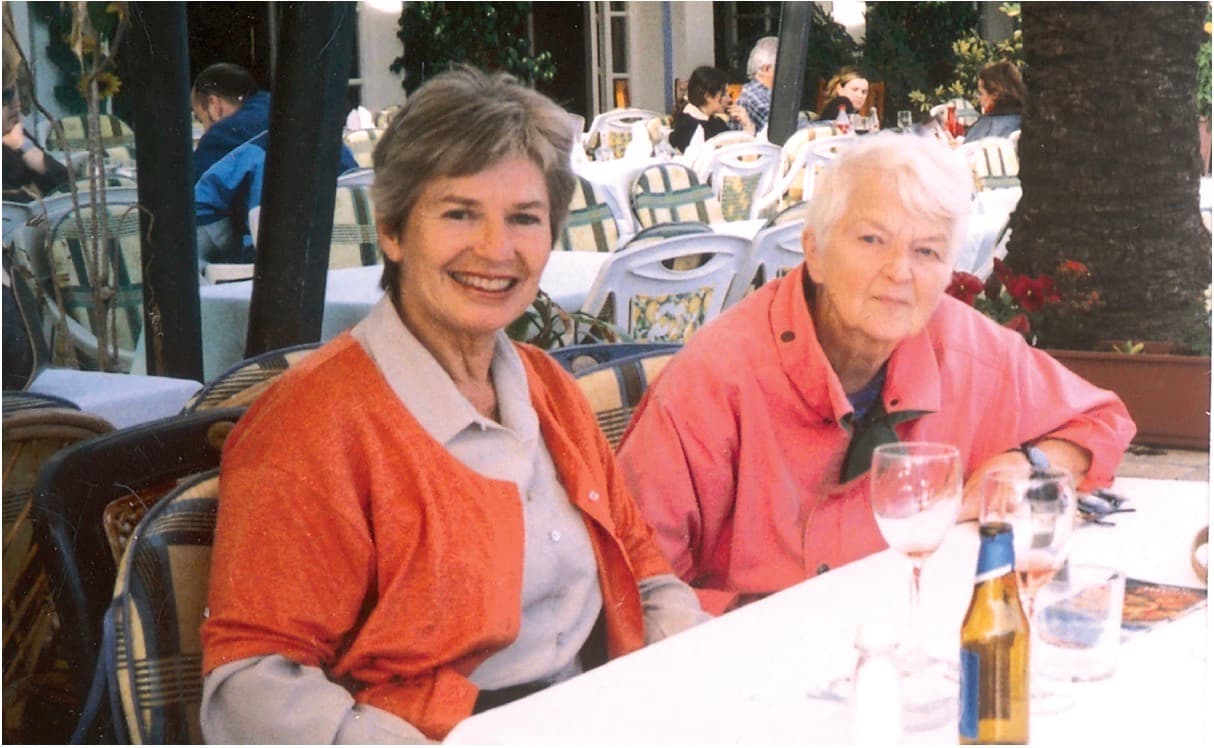 Fargefotografi av to eldre kvinner som sitter ved et bord på en uteservering i solskinn.