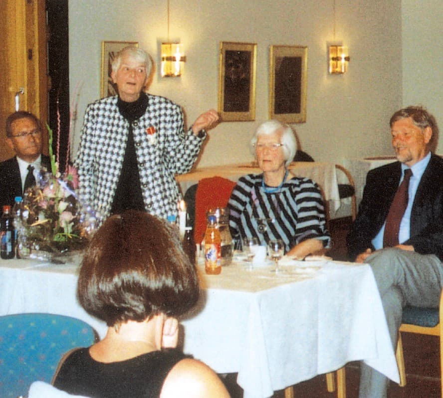 Fotografi fra en tilstelning. En kvinne i mønstret jakke står og taler ved et bord med gjester rundt.