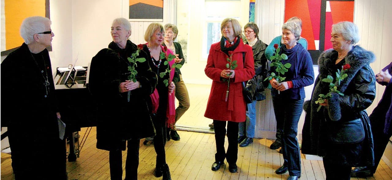 Fotografi fra en utstillingsåpning. En gruppe kvinner står med røde roser i hendene i et galleri med billedtepper på veggene.