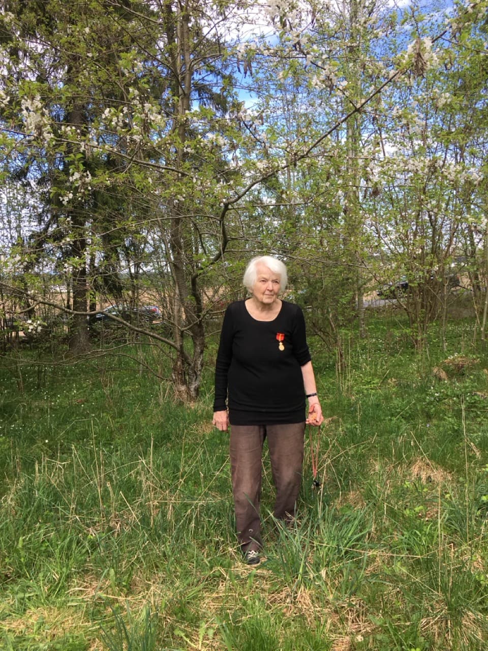 Fargefotografi. Karin står i høyt gress under blomstrende trær, i svart genser med en medalje i rødt bånd på brystet.